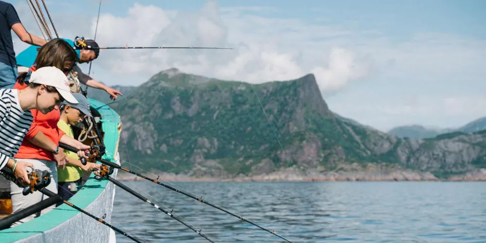 Guests fishing from the boat railing beneath dramatic Lofoten peaks